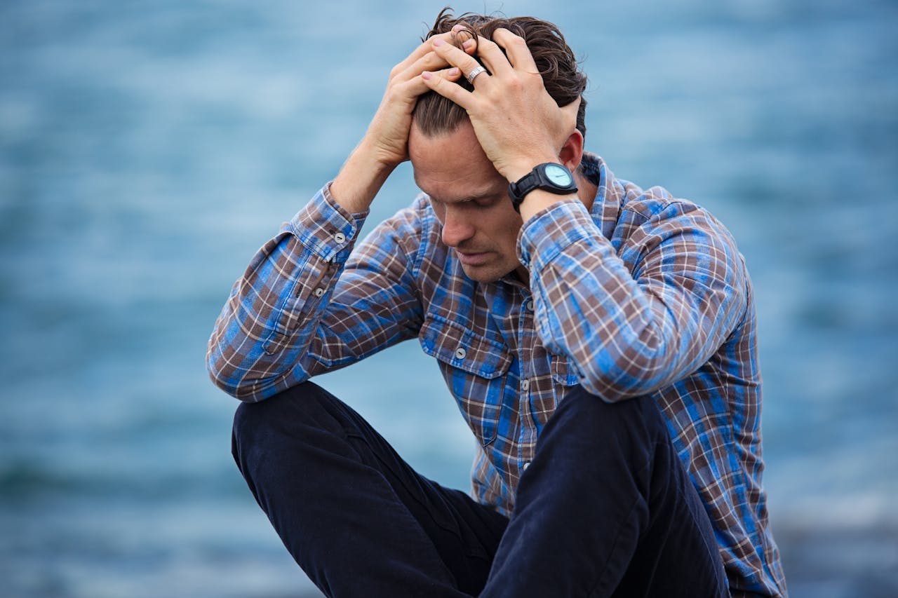 A man in a plaid shirt sits by the water looking depressed, symbolizing stress.