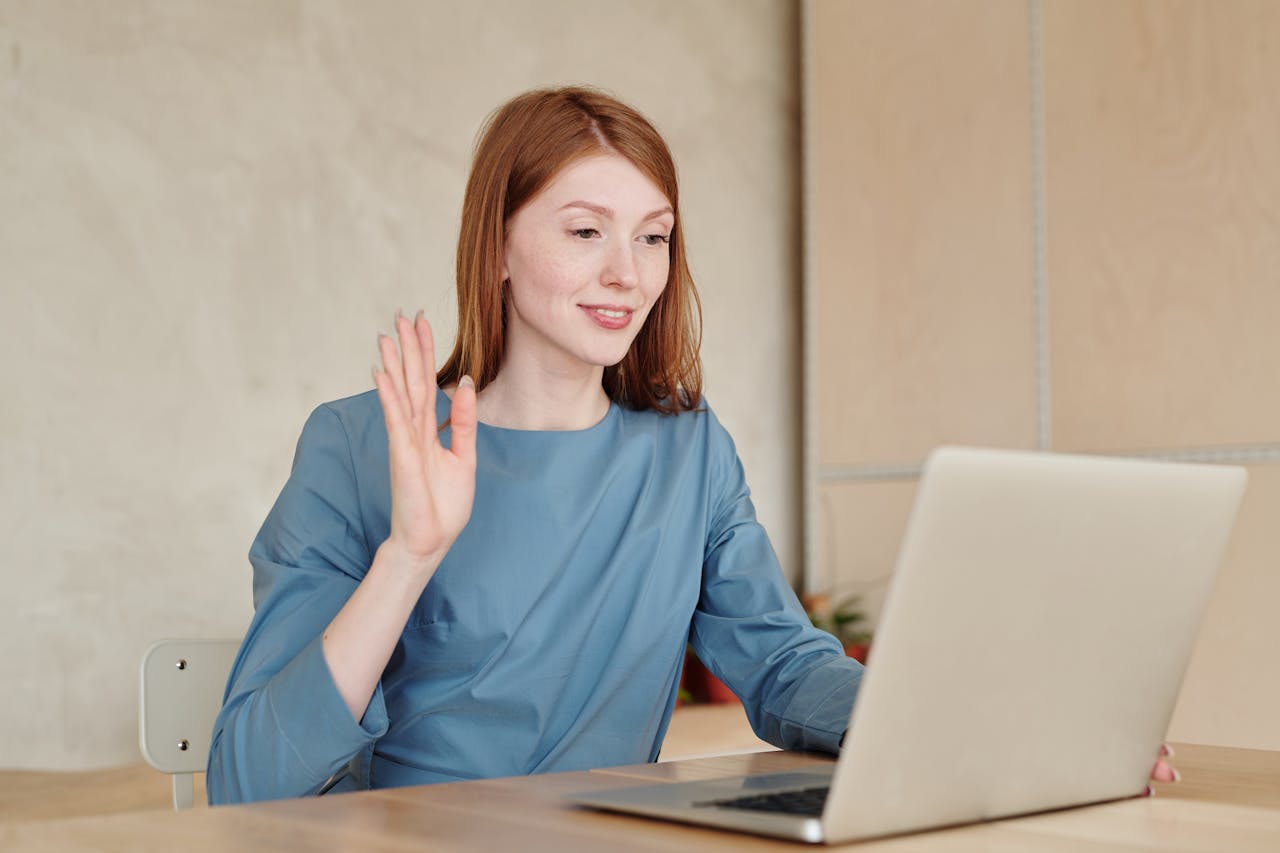 Smiling woman in blue shirt waving during a video call on her laptop.